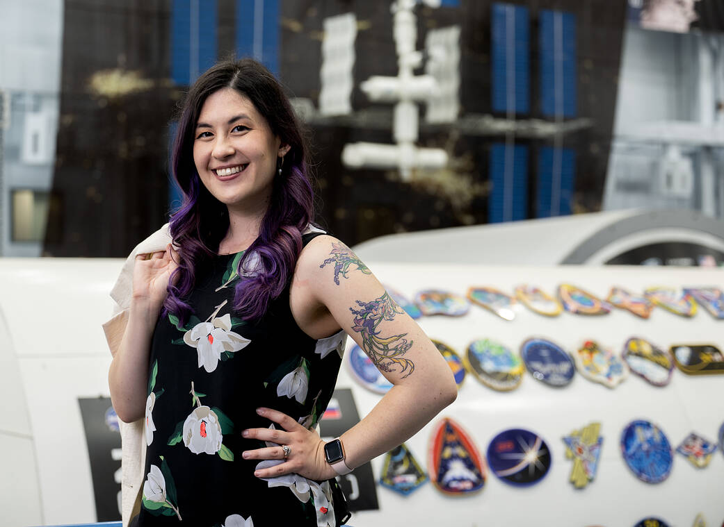 ISS Research Communications Lead Jennifer Hamilton poses and smiles at the camera in the Mockup Facility at NASA's Johnson Space Center. She is carrying a light pink blazer over her shoulder.