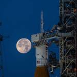 A full Moon is seen in the night sky at center left with the top of the Space Launch System rocket and Orion spacecraft upright in the center of the frame. The mobile launcher is visible in the sides of the frame.