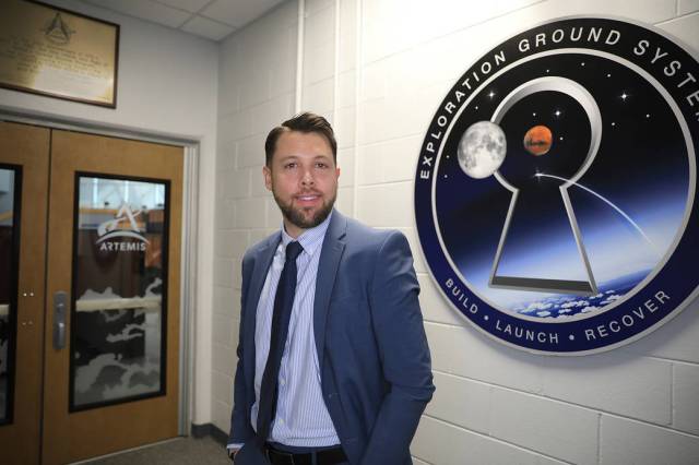 Social Media Specialist Chad Siwik stands in front of a doorway at NASA's Kennedy Space Center. He is wearing a suit and tie and is smiling at the camera.
