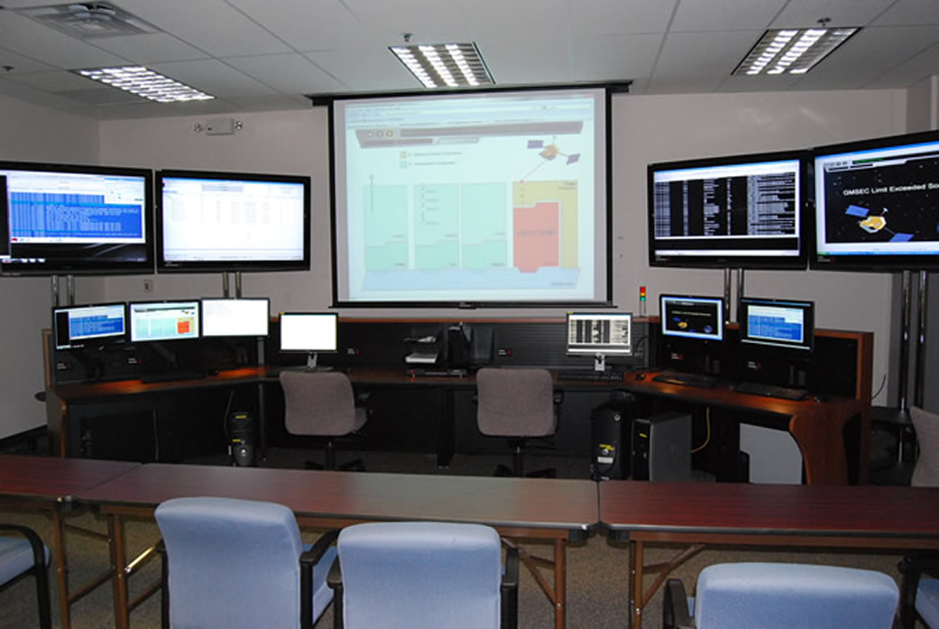 A room with a large projector screen in the back with other monitors on the wall and rows of tables.