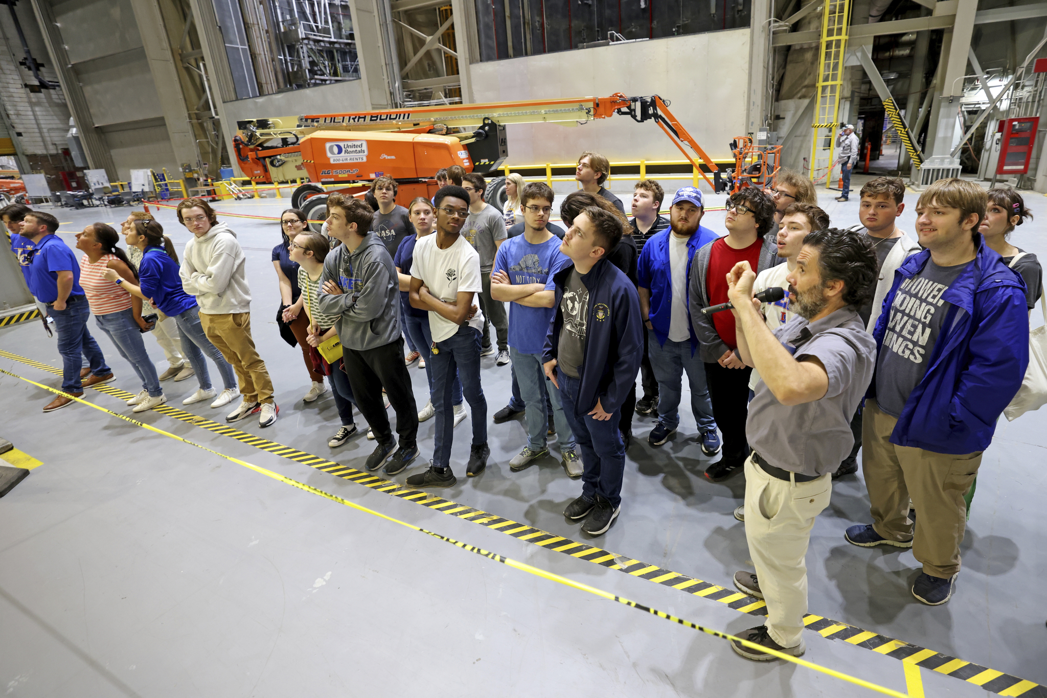 A group of students stand on a concrete floor behind yellow and black striped tape on the ground as a employee gives a tour with a microphone.