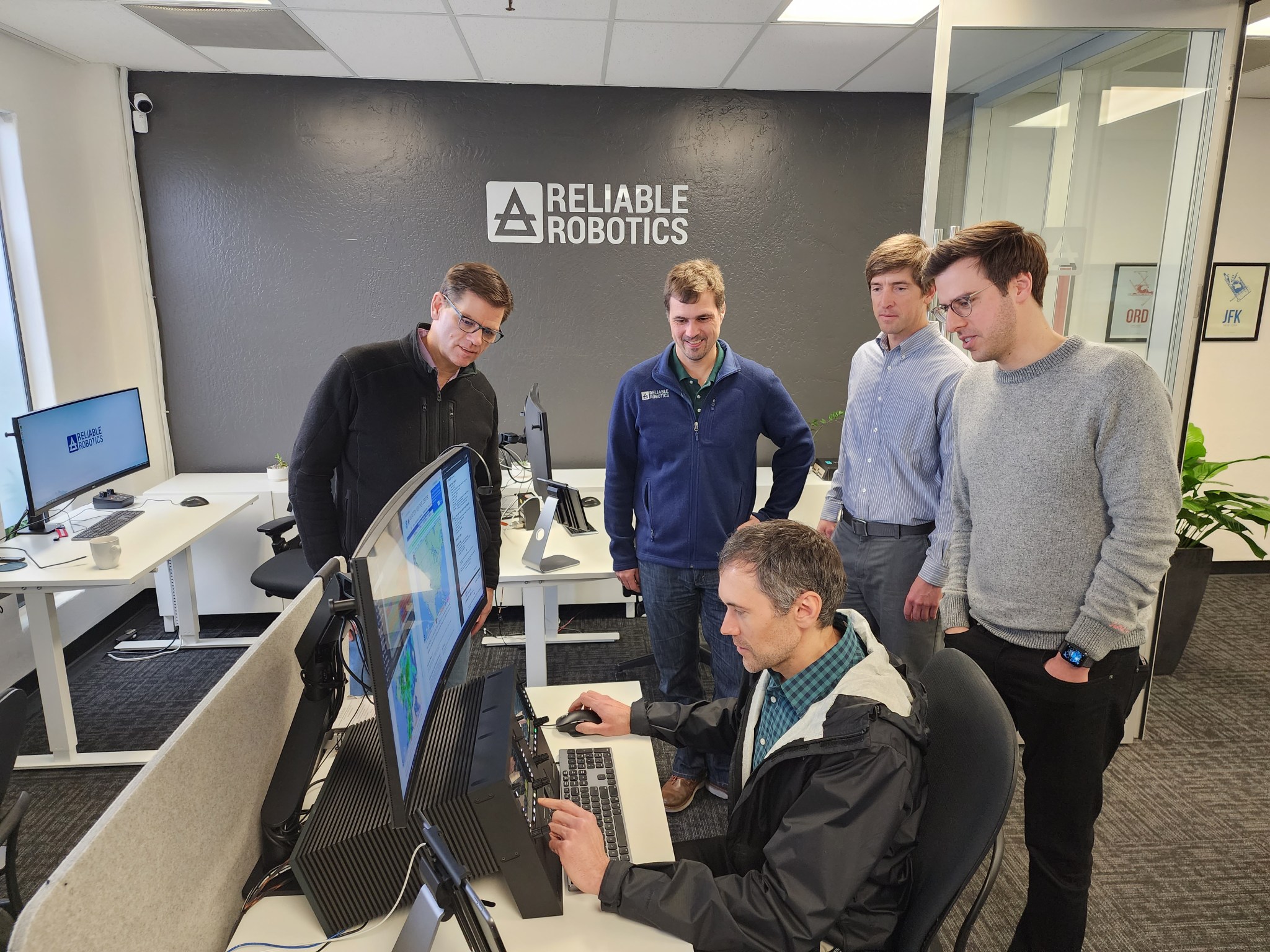 Four people stand around one person who is sitting behind a computer.