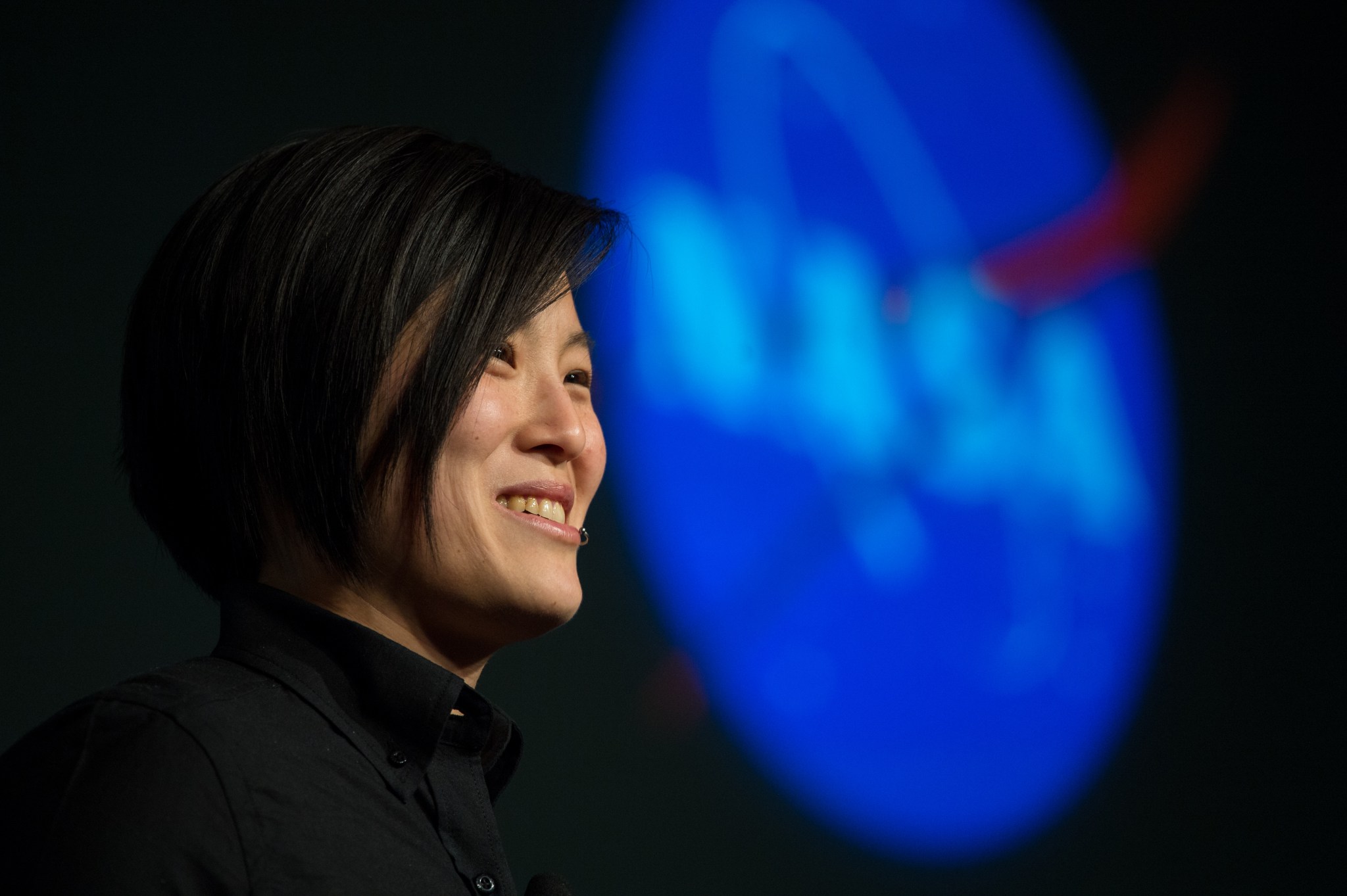 Tina Lai stands in front of a lighted NASA insignia speaking to an unseen group of people