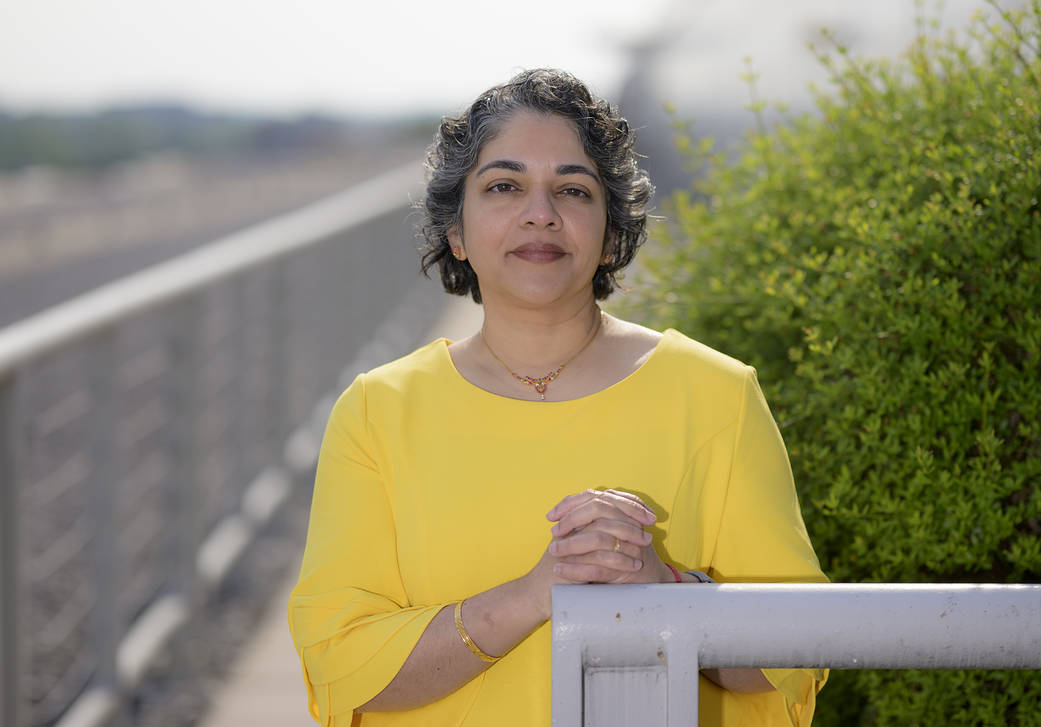 An Indian woman with short, curly, salt and pepper hair smiles softly at the camera wearing a sunshine yellow quarter sleeve blouse. Her hands are clasped as she leans agains a railing outside, with bright green bushes on her right-hand side.