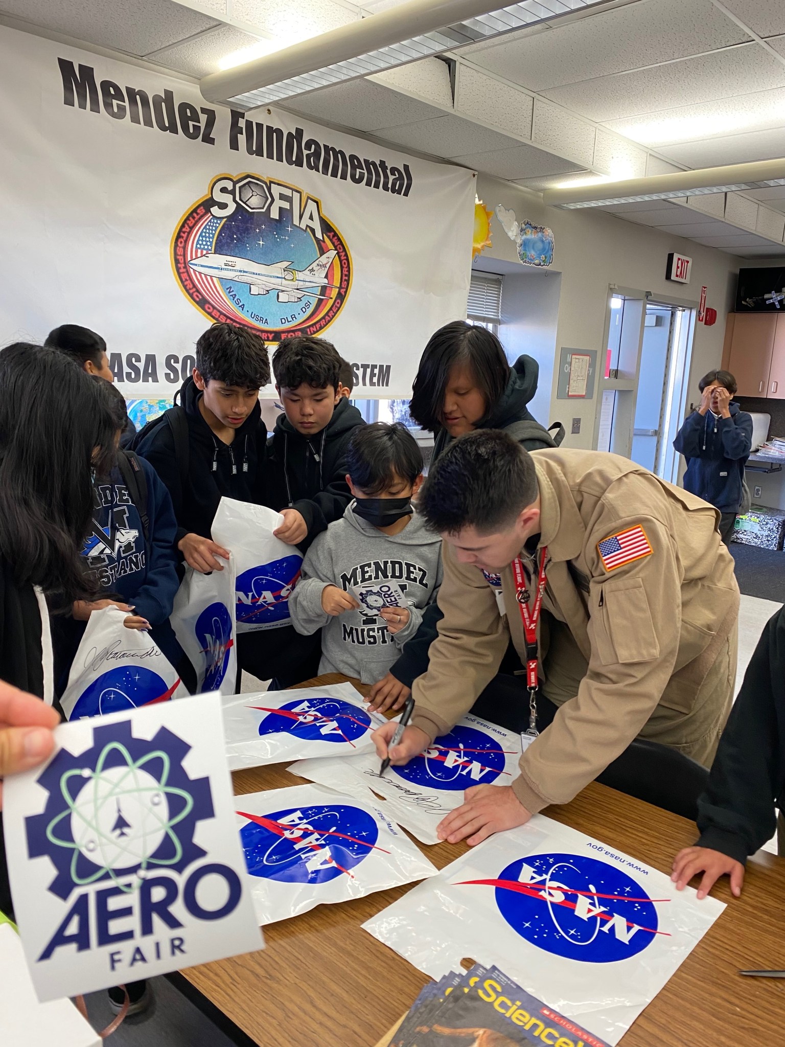 Students gather around with shinning bags with NASA printed on them, while an adult signs the bags.