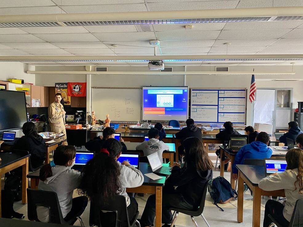 Adult stands at the front of a classroom pointing at a digital screen, students sit at desks, some with laptop open.