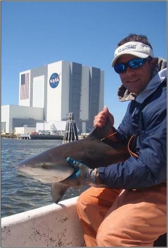 A man is photographed holding a bull shark at NASA's Kennedy Space Center in Florida. In the background is the iconic Vehicle Assembly Building.