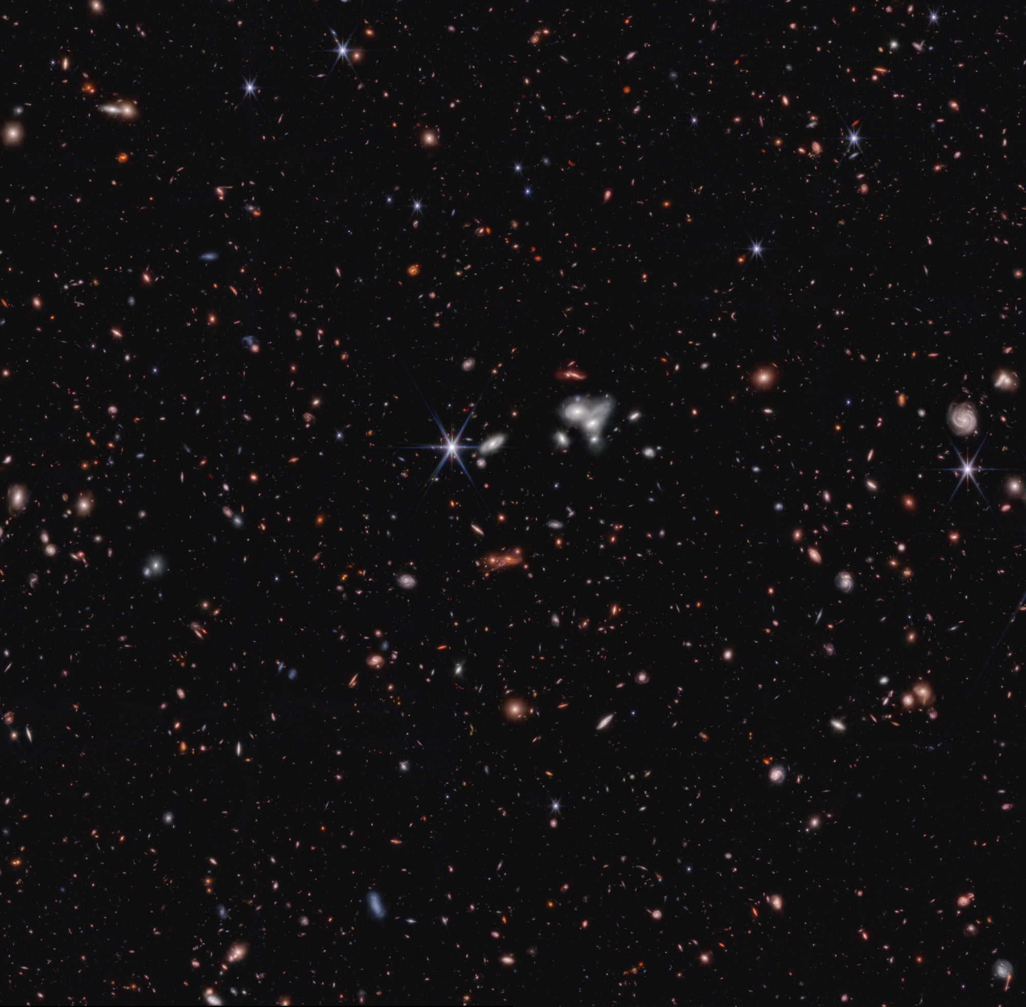 About 100,000 galaxies appear with many overlapping objects at various distances. They include large, blue foreground stars, some with all eight diffraction spikes, white and pink spiral and elliptical galaxies, and many tiny red dots throughout.