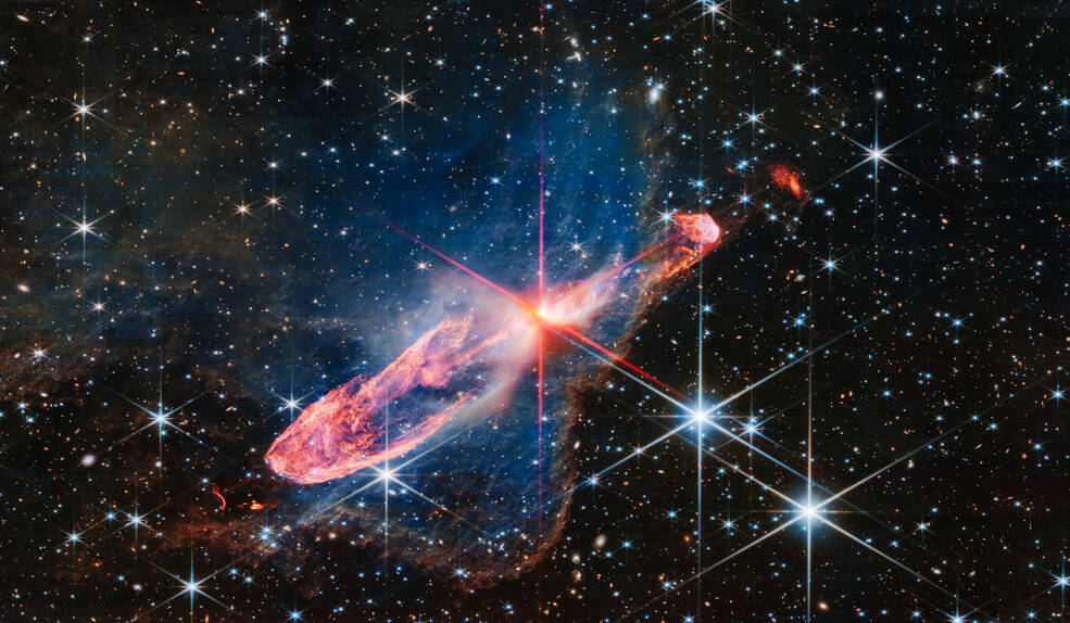 Horizontal orange cloud known as Herbig-Haro 46/47, with a central yellow-white blob pierced by red diffraction spikes. A surrounding nebula is seen as a delicate, semi-transparent blue haze. The background is filled with stars and galaxies.