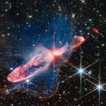 Horizontal orange cloud known as Herbig-Haro 46/47, with a central yellow-white blob pierced by red diffraction spikes. A surrounding nebula is seen as a delicate, semi-transparent blue haze. The background is filled with stars and galaxies.