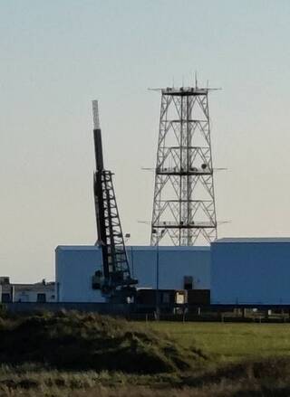 a rocket sits on a pad at a slight angle to the left under a clear sky with a low blue building and a tower in the distance.