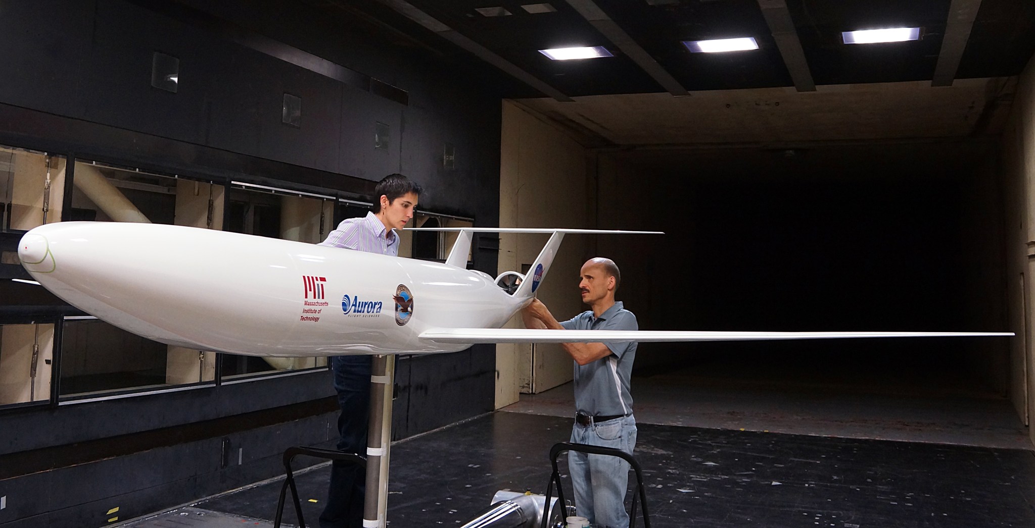 Two people work on a scale model of an airplane inside a wind tunnel.
