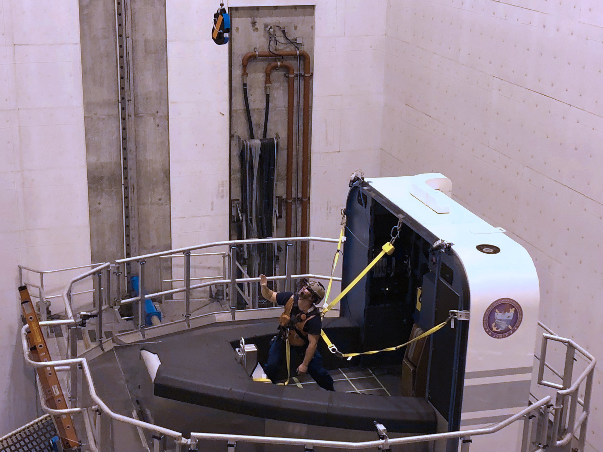 A human participant is shown attached to a simulation cab, resembling a small boat, on the VMS. The participant is looking and reaching up.