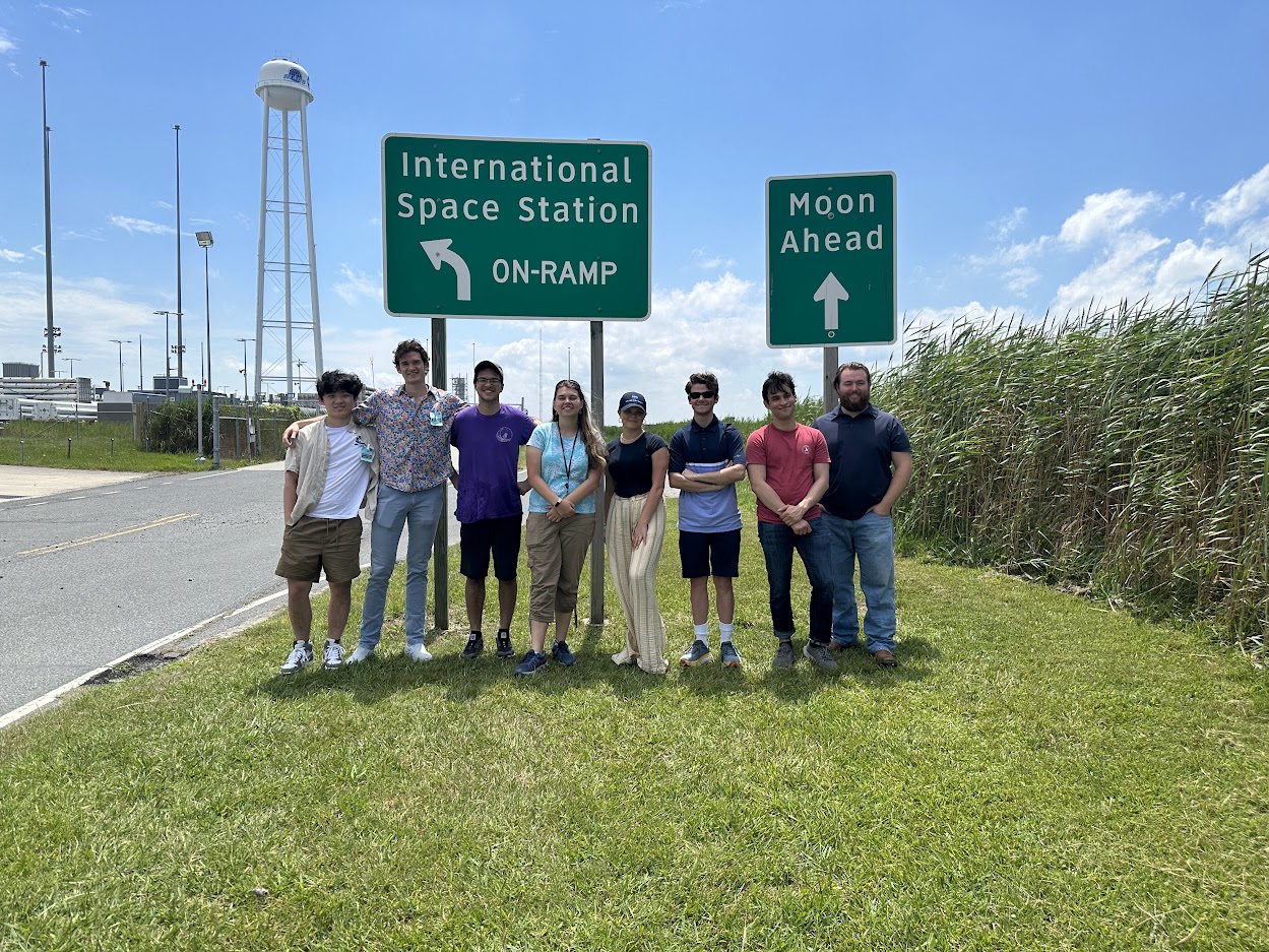 A group of summer interns at Wallops Flight Facility smiling in front of two traffic direction signs that state: International Space Station and The Moon.