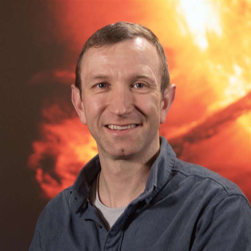 A man, seen from the shoulders up. He is smiling and standing in front of an image showing a portion of the Sun.