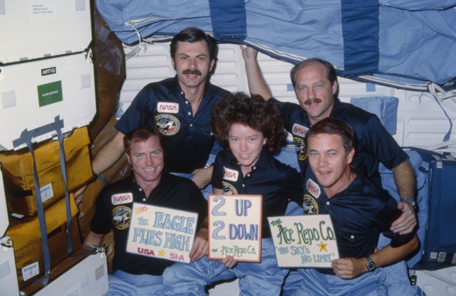 The five-member 51-A crew celebrates a successful mission. The front row of astronauts are holding up signs which say "The Eagle Flies High", "2 up, 2 down, the Ace Repo Co." and "The Ace Repo Co., The Sky's no limit". The reference to the Eagle has to do with the Discovery crew's mascot, which appeared both in the crew portrait and insignia.