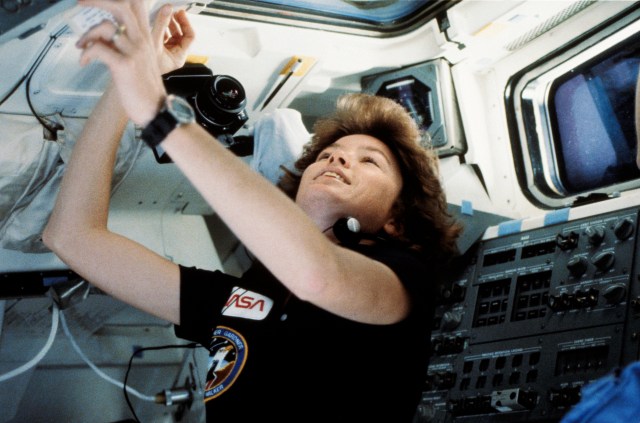 Astronaut Anna L. Fisher is pictured near the aft flight deck of Discovery, where she appears to be taking photos from the observation station. A camera floats just above her head.