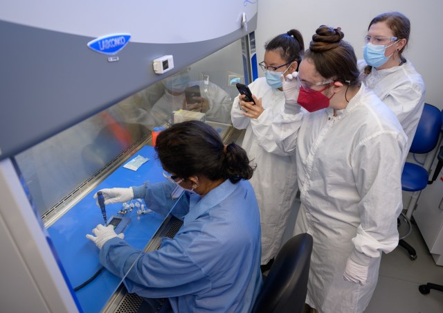 A person uses a tool to connect electronics to a manifold while three researchers observe.