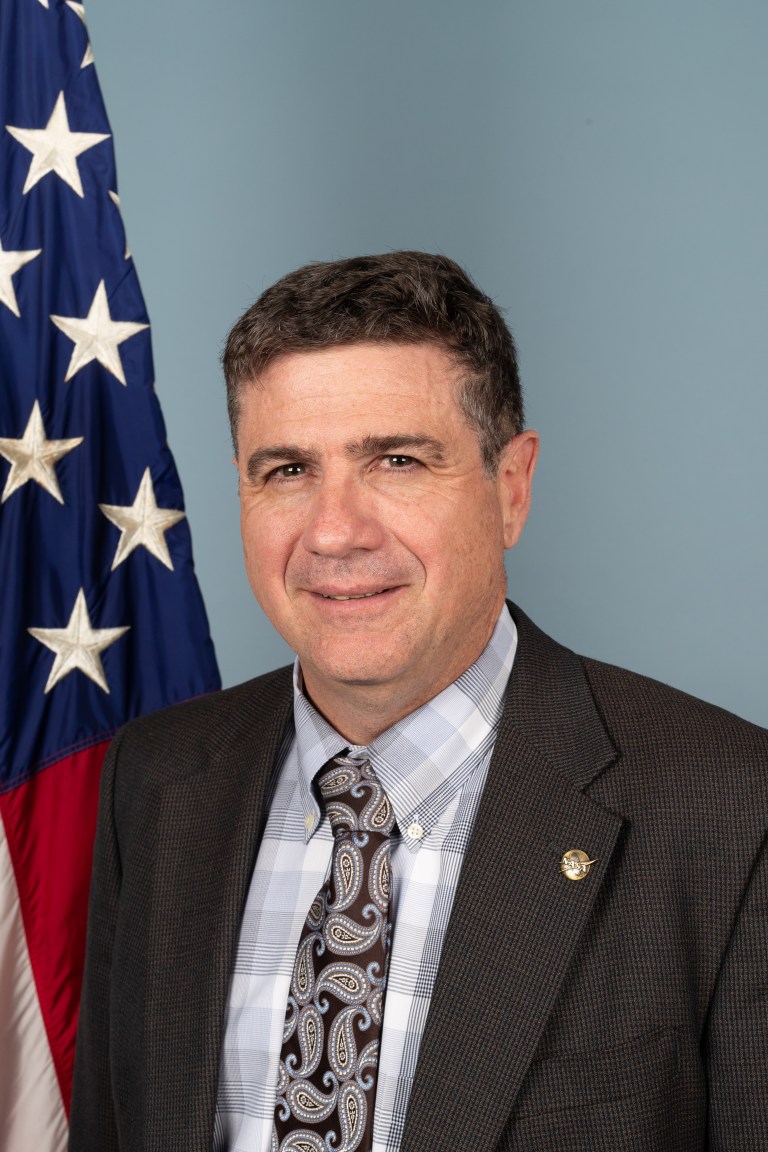 Robert Jameson poses for a portrait in front of an American flag against a blue background.
