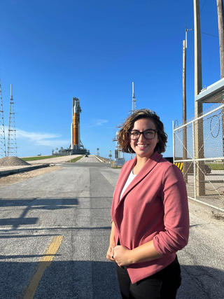 Darcy DeAngelis poses at NASAs Kennedy Space Center. Artemis I can be seen in the background on the launchpad. The weather is sunny, and the sky is blue. DeAngelis is wearing professional attire.