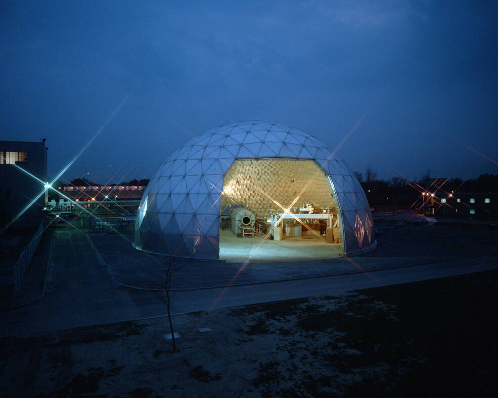 A white, half-dome shaped building with triangular sections on its exterior is illuminated at nighttime on NASA Glenns campus. Through the facilitys open door, test rigs can be seen inside.