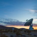 A large antenna, part of the Deep Space Network, sits on top of a hill at sunset. The sky around the antenna is a bright orange but bleeds into a dark blue around the edges of the image.