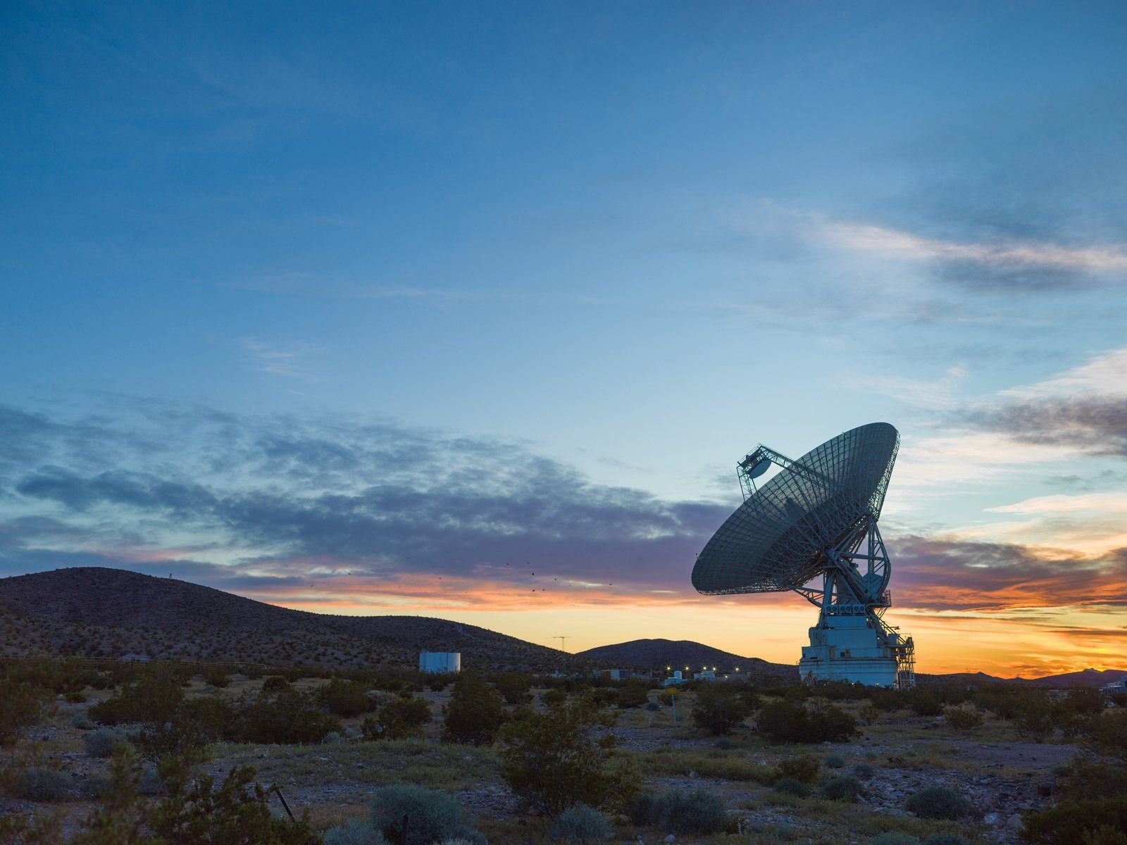 A large antenna, part of the Deep Space Network, sits on top of a hill at sunset. The sky around the antenna is a bright orange but bleeds into a dark blue around the edges of the image.