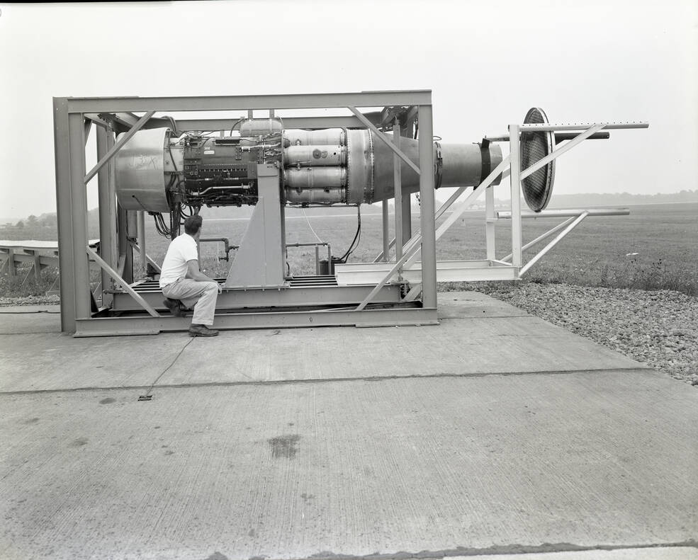 A black-and-white image of a metal frame holding up a jet engine outside at NASA Glenn. A researcher inspects the frame, sitting below the engine.