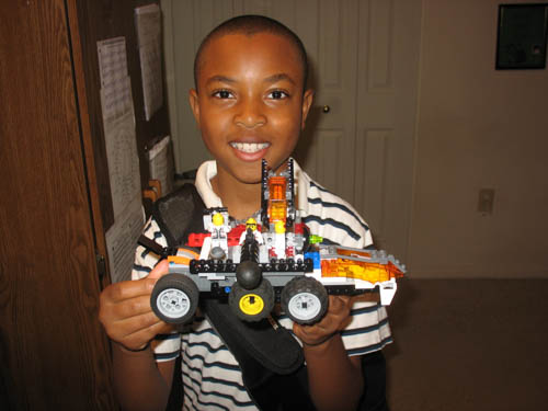 A small boy smiles and holds a LEGO car with a proud smile. He stands in front of a tall brown cabinet in his home.