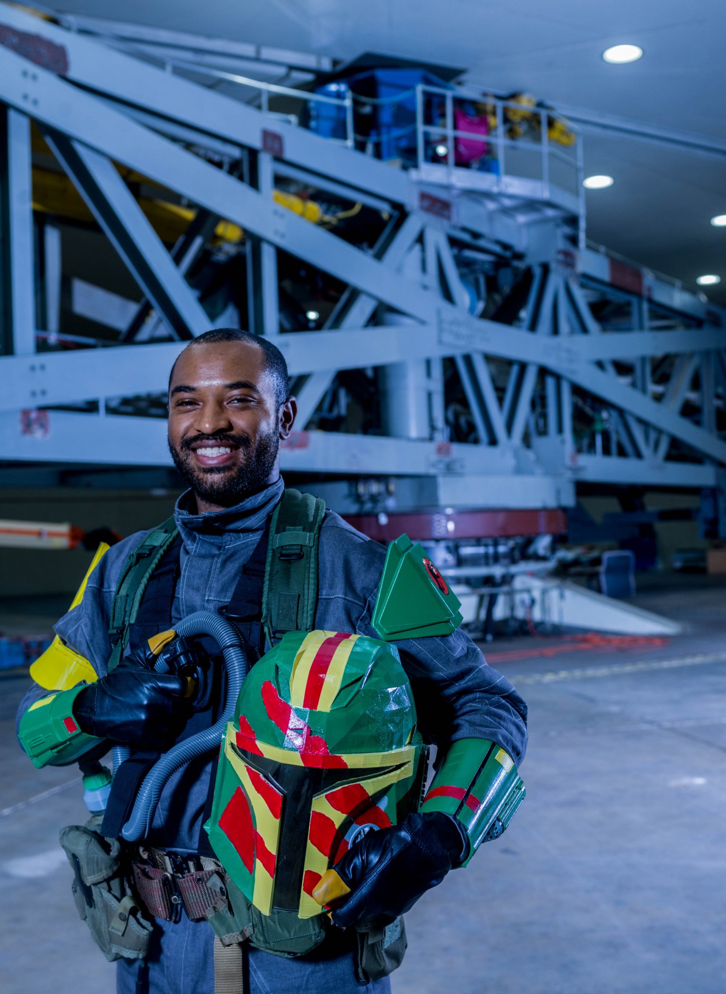 A man in a colorful Star Wars-style Mandalorian costume smiles and poses in front of Goddard's centrifuge, a huge room with a horizontal metal framework in the center.