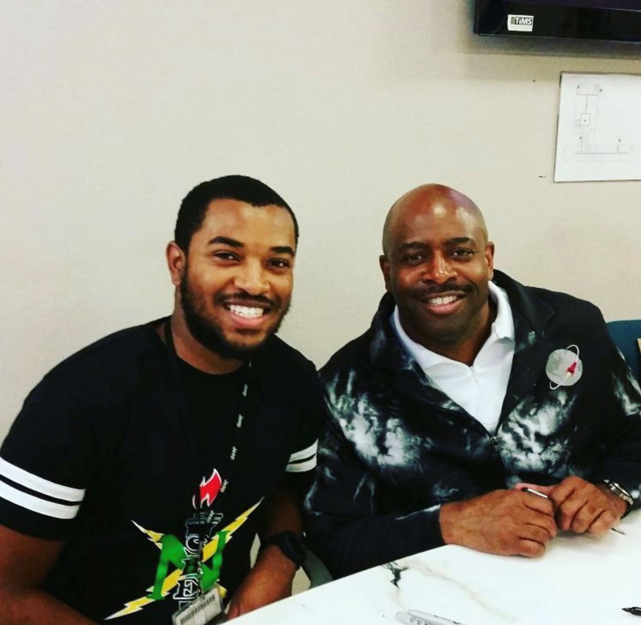 Two men sitting at a table smile at the camera in an informal snapshot. The younger man on the left wears an honor society T-shirt. The older man on the right wears a jacket with an orbiting rocket logo.