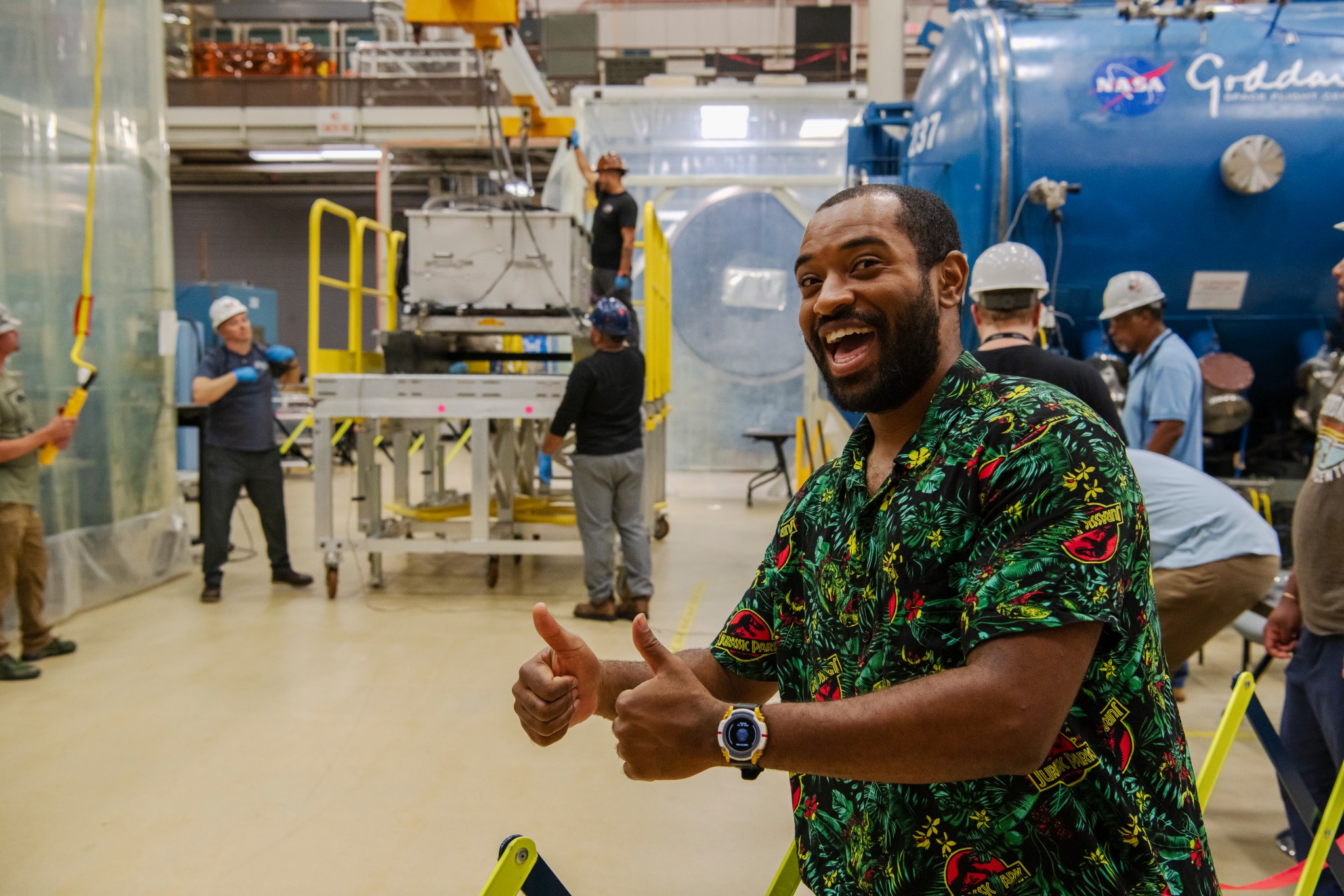 A man wearing a colorful jungle-print shirt grins and poses with two thumbs up in Goddard's clean room. Other employees in work clothes and hardhats work in the background.