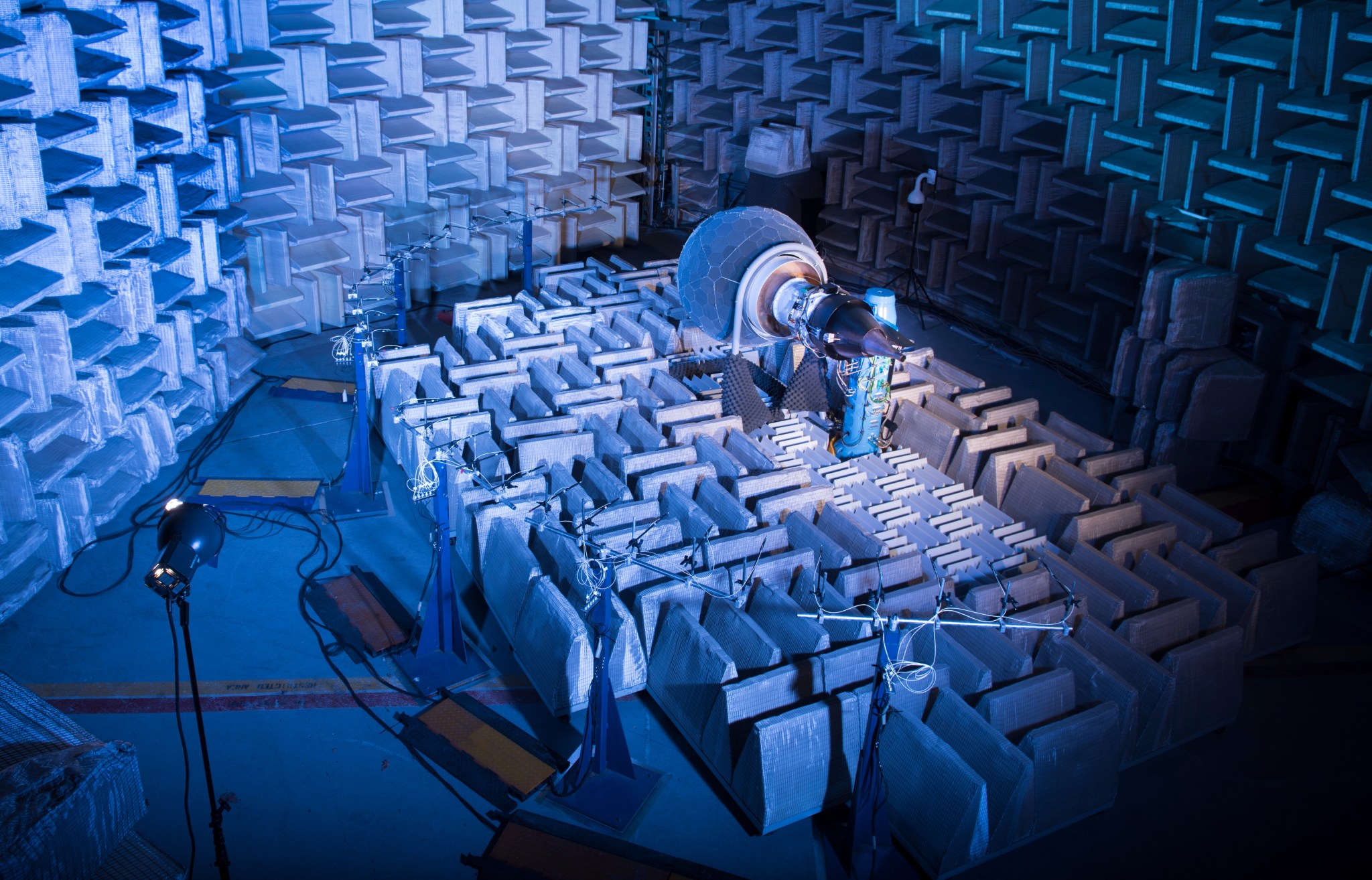 The DGEN AeroPropulsion Research Turbofan test rig is illuminated by lights inside of NASA Glenn’s Aero-Acoustic Propulsion Laboratory. The walls of the facility, covered in wedges, glow blue in the background.