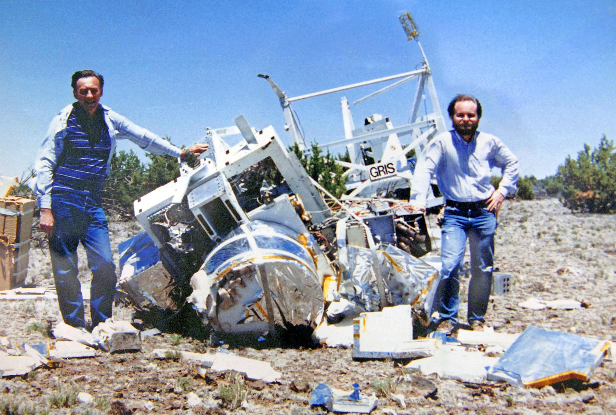 Two men flank a pile of white and silver wreckage on which the word "GRIS" is visible.