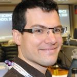 Photo of man with dark brown hair, wearing glasses, smirking at the camera in a brown collared shirt.