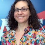 Portrait of a woman with curly black hair, smiling at the camera wearing black glasses, wearing a colorful button-up shirt with light blue, orange, magenta, yellow, and white flowers.