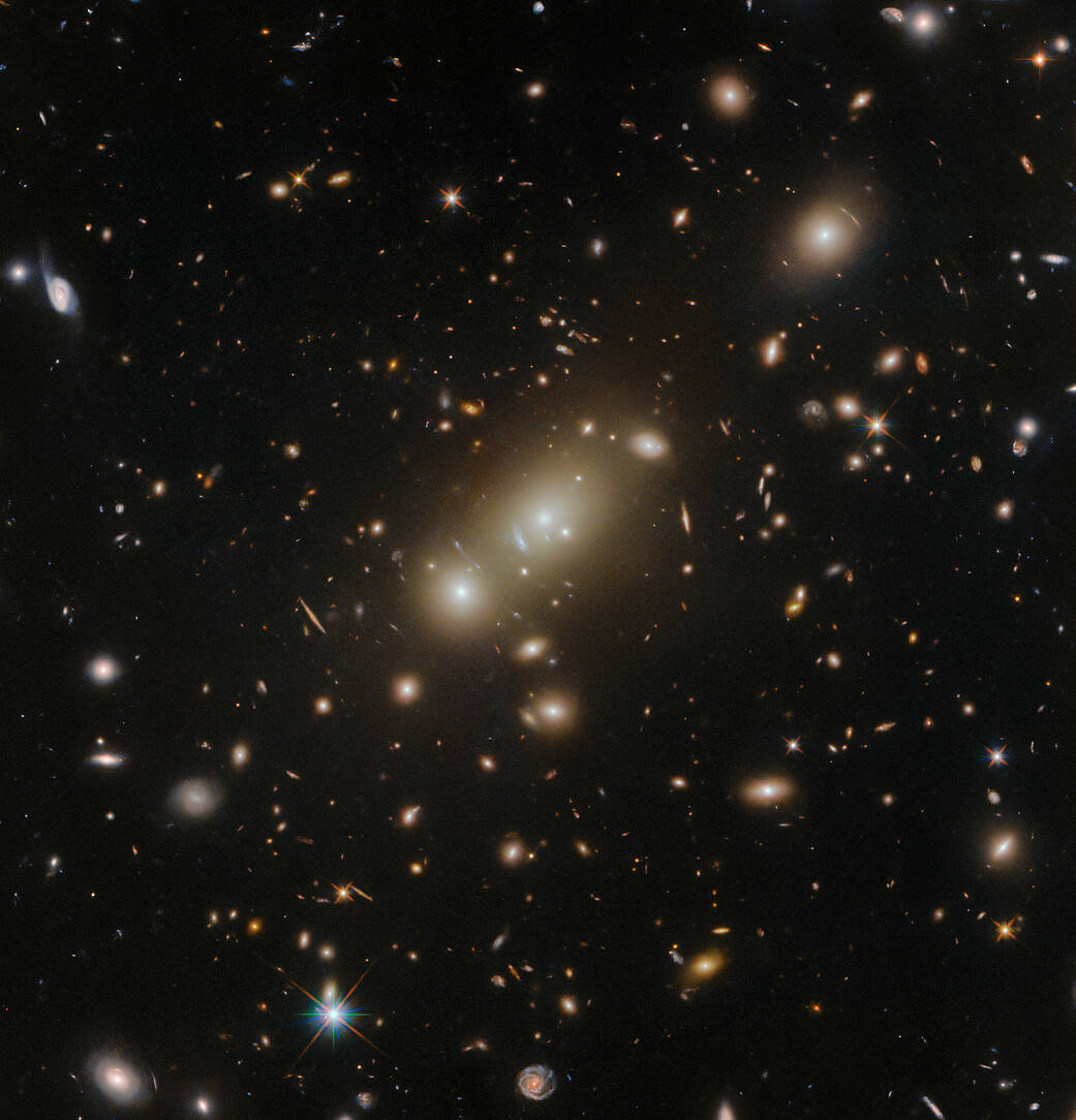 A crowd of oval-shaped elliptical galaxies gather around the largest galaxy in the center. They are surrounded by more distant stars and galaxies, that have many shapes and sizes but all are smaller, on a dark background.