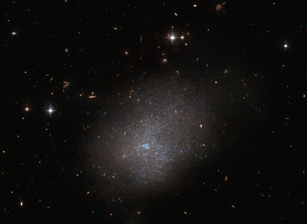 Irregular galaxy of many tiny stars clumped together, surrounded in diffuse light with a bubble of blue gas in its bright center. It is surrounded by mostly small, faint objects with a few bright stars above and left of it and a string of galaxies nearby.