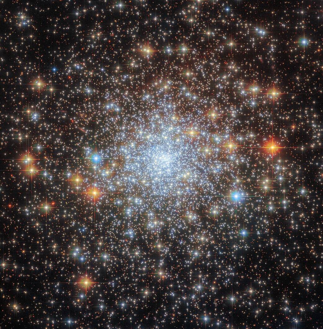 A spherical cluster of stars with a bright core, and stars spread out to the edges gradually giving way to an empty, dark background. A few stars with cross-shaped diffraction spikes appear larger and stand out in front.