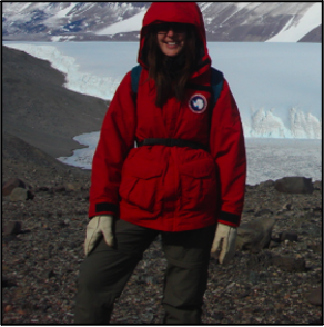 Jennifer Heldmann wearing a red heavy winter jacket and white gloves and sunglasses. Standing in front of a a cold body of water with ice and white and brown mountains behind.