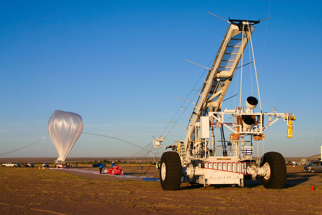 A scientific balloon is to the left, and appears as a plastic, upside down teardrop. A tube attached to the top of the balloon leads down to the ground. A crane to the right holds a large payload structure.