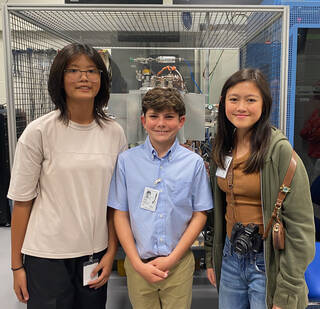 Three young students wearing name badges pose in front of a piece of hardware inside NASA Glenns Stirling Research Laboratory.