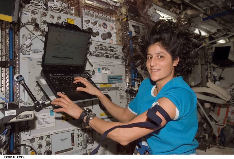 Image of NASA astronaut Sunita Williams looking toward the camera and smiling as her hands are on the keyboard of a laptop computer attached via Velcro to a flat surface connected to a rack on the Space Station.