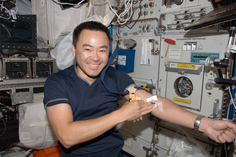 Image of JAXA astronaut Akihiko Hoshide looking toward the camera as he uses his right hand to hold an absorbent pad on his extended left arm. His right hand also holds several vials of blood from the blood draw that he has just completed.
