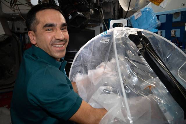 Image of NASA astronaut Raja Chari looking toward the camera as his hands manipulate objects in a portable glove bag for the Food Physiology investigation.