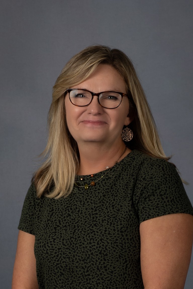 A blonde woman with glasses in a black shirt sits in front of a grey background for her headshot.
