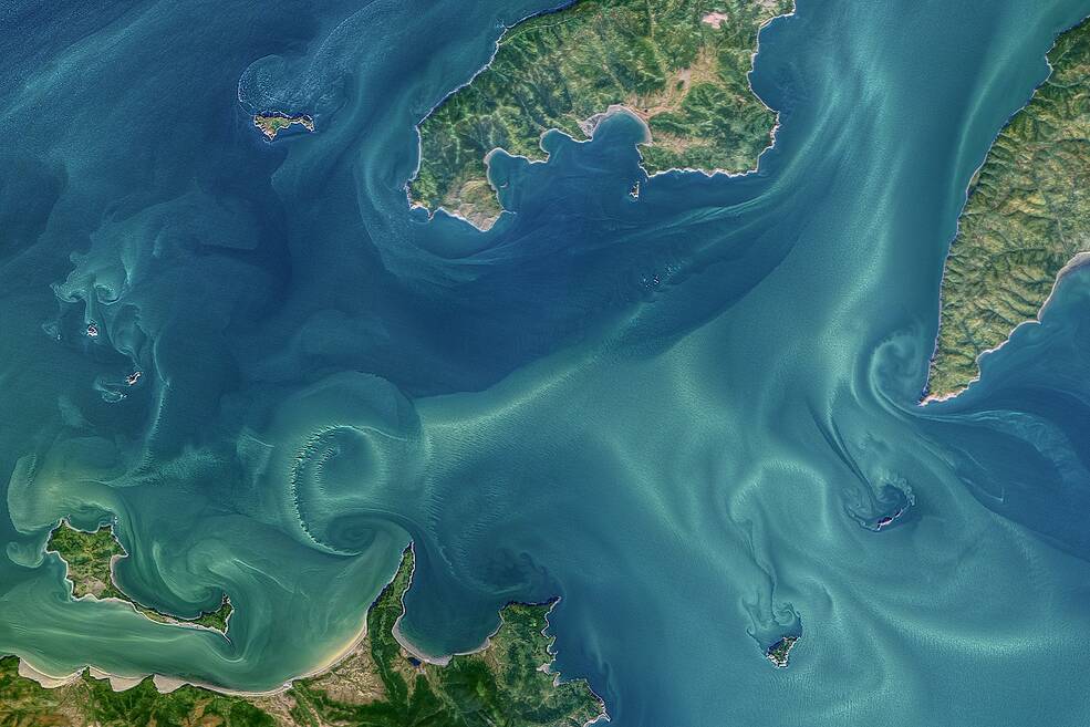 A satellite image of tide currents with Blue water with white swirls and green land mass. The region shown includes some of the Shantar Islands and part of Uda Bay in the western Sea of Okhotsk.