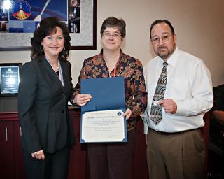 Three people in professional clothing pose in an indoor space with tan walls. The person in the middle, Barbara McKissock, holds an award certificate, and the person on the right holds a pin.