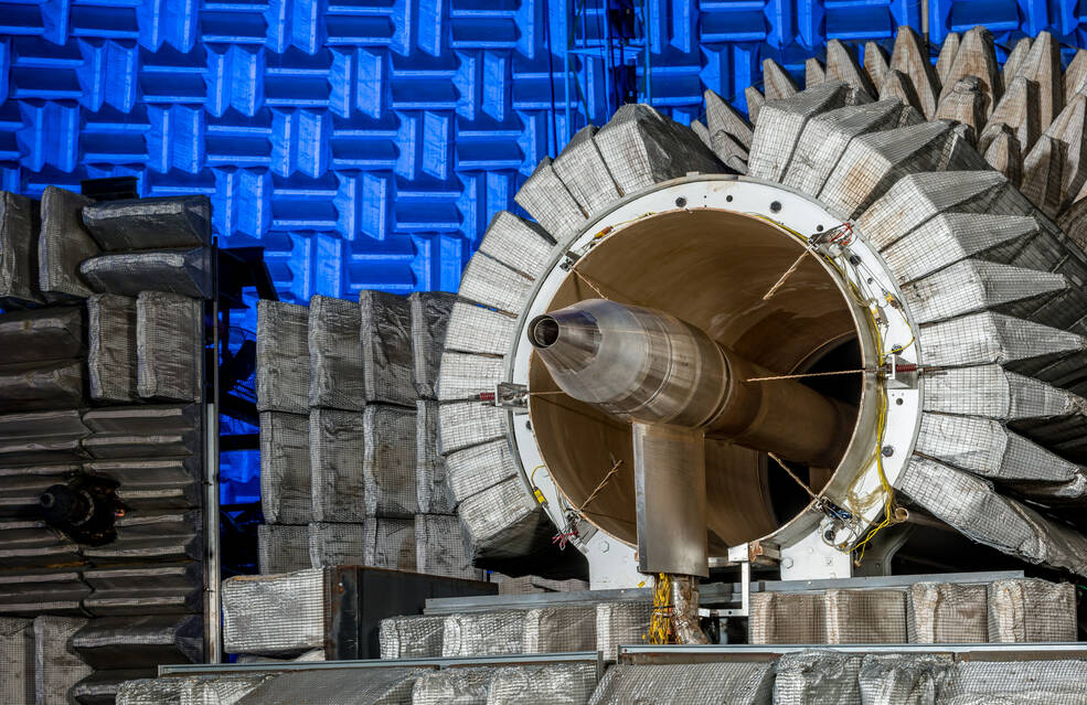 A Learjet engine nozzle peeks out from NASA Glenns Nozzle Acoustic Test Rig, a cylinder surrounded by tan wedges. The walls of the Aero-Acoustic Propulsion Facility, also covered in wedges, glow blue in the background.