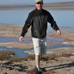 Richard Craig Everroad at what appears to be a marsh wearing sunglasses, a tan hat, black jacket, and tan cargo pants with black wet shoes on.